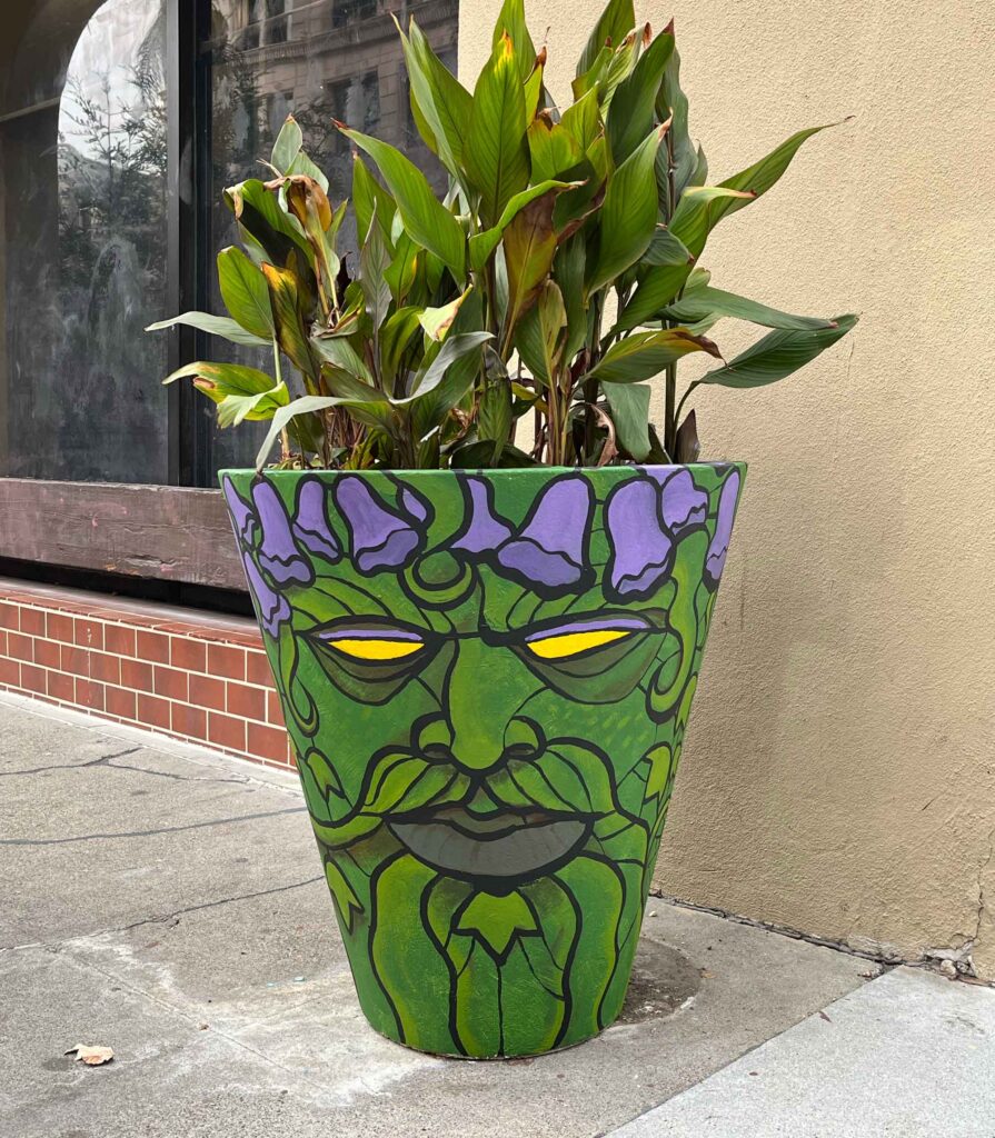 Green Man of South First, a waist-high conical planter painted as a mustached green man wearing a crown of jacarinda with yellow glowing eyes